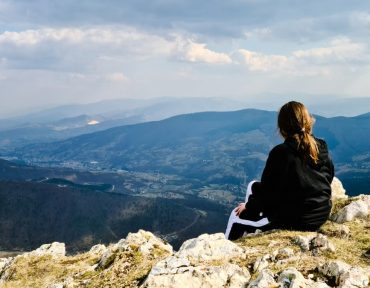 A woman sits on a rocky mountain peak, enjoying a panoramic view of the valley below.