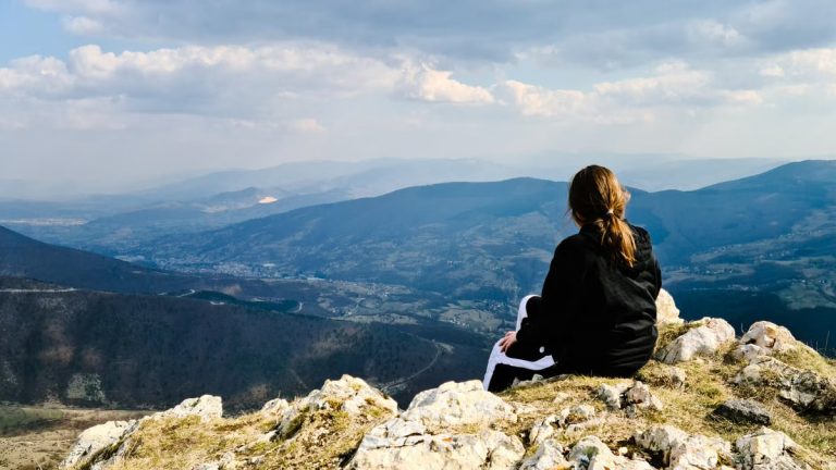 A woman sits on a rocky mountain peak, enjoying a panoramic view of the valley below.